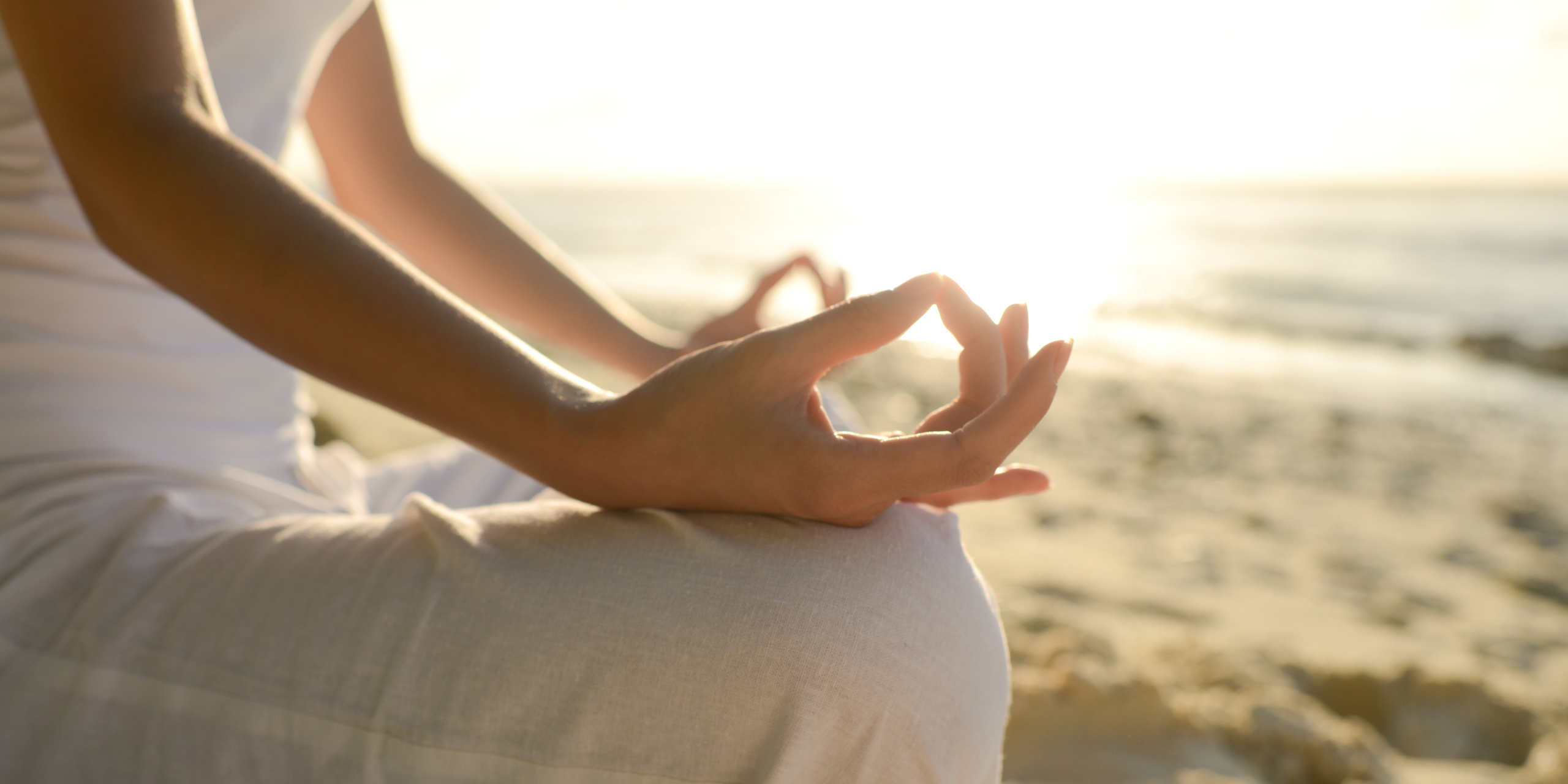 lady in white sitting on abeach sub riseing in background yoga meditation